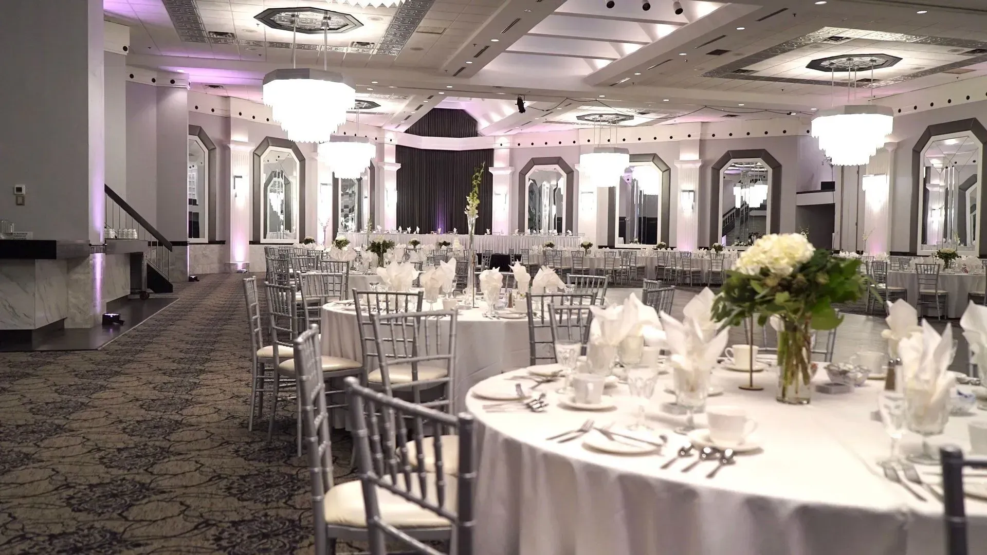 Elegant banquet hall set with round tables, white tablecloths, silver chairs, and floral centerpieces.