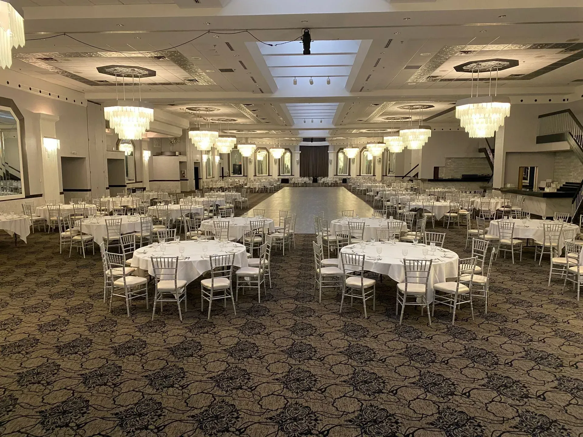 Large banquet hall with round tables and white chairs, set for an event under soft ceiling lights.