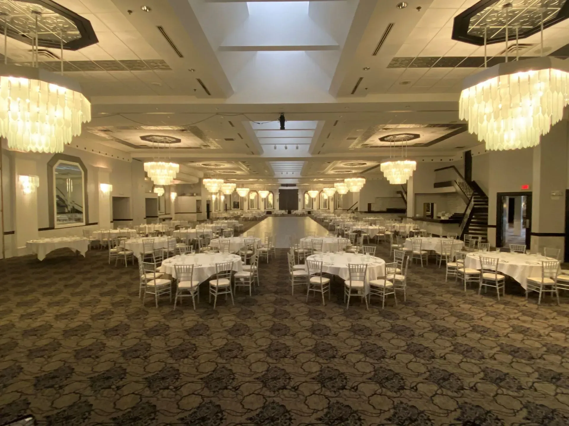 Elegant banquet hall with round tables, white chairs, chandeliers, and patterned carpet