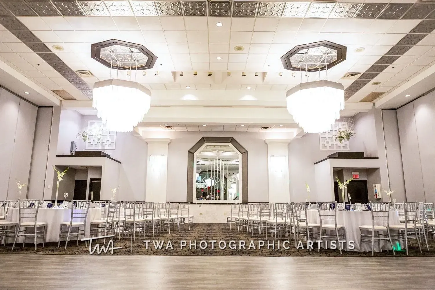 Elegant banquet hall with chandeliers, mirrored wall, and rows of silver chairs and tables