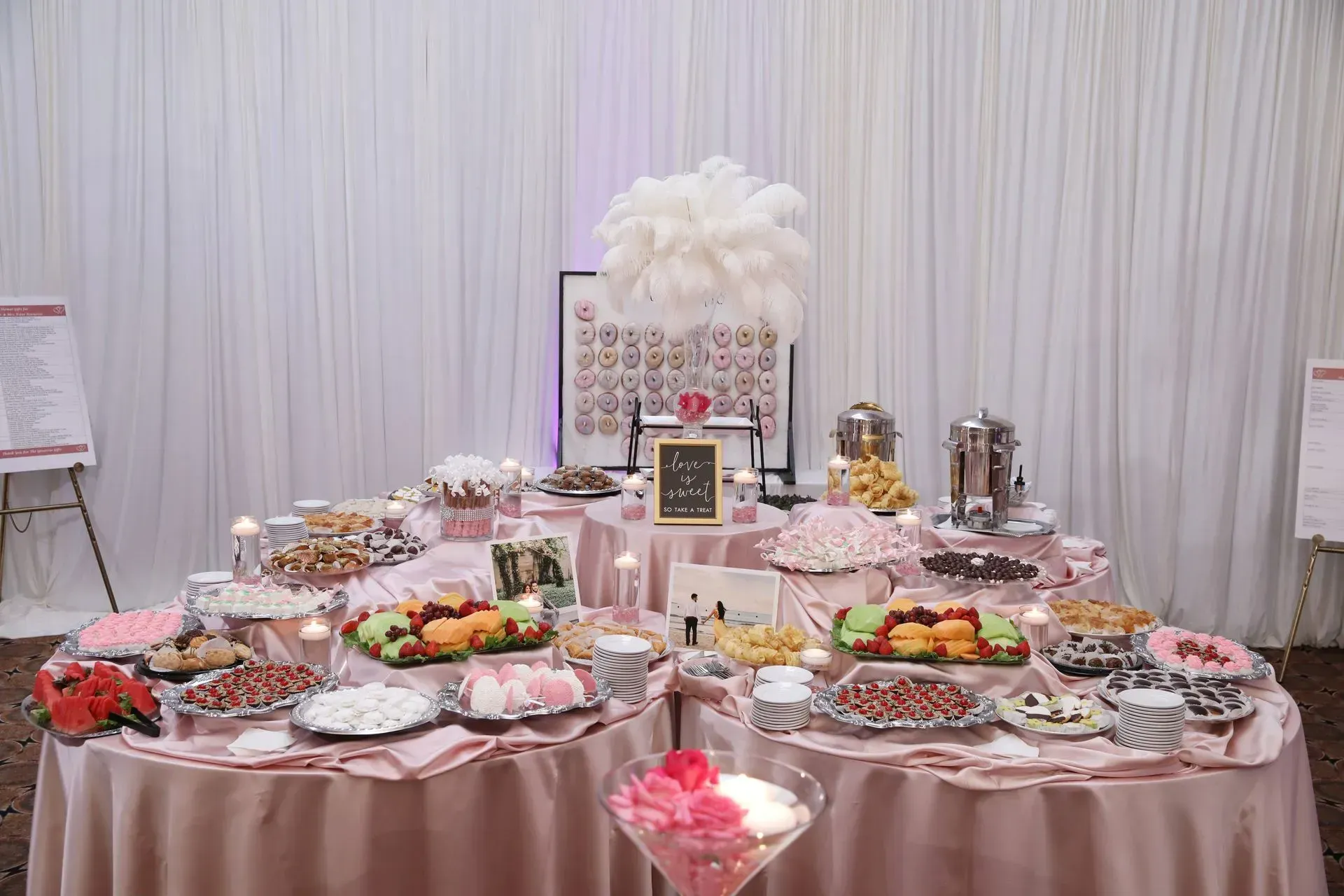 Buffet table with assorted desserts, fruit, and drinks, decorated in white and pink for an event
