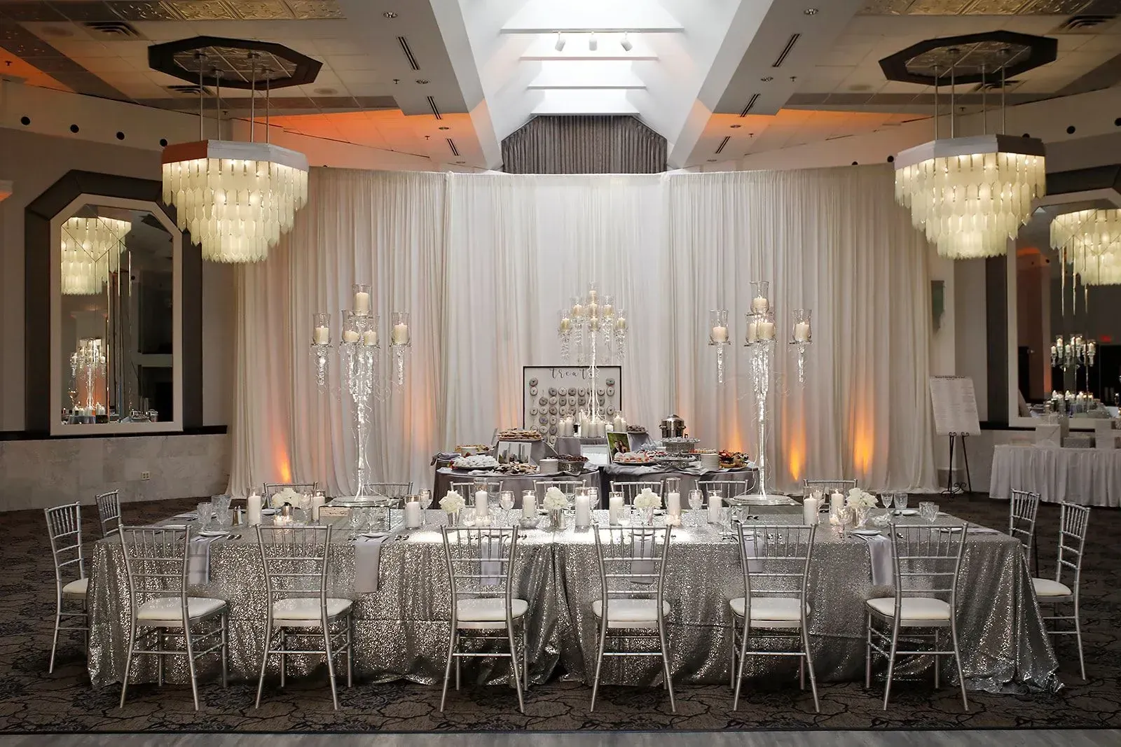 Elegant banquet hall with silver chairs, white drapery, chandeliers, and a decorated central head table