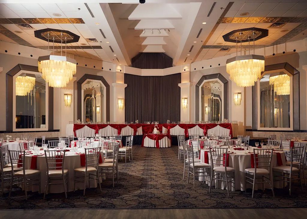 Elegant banquet hall with round tables, red chairs, chandeliers, and a dark patterned carpet.