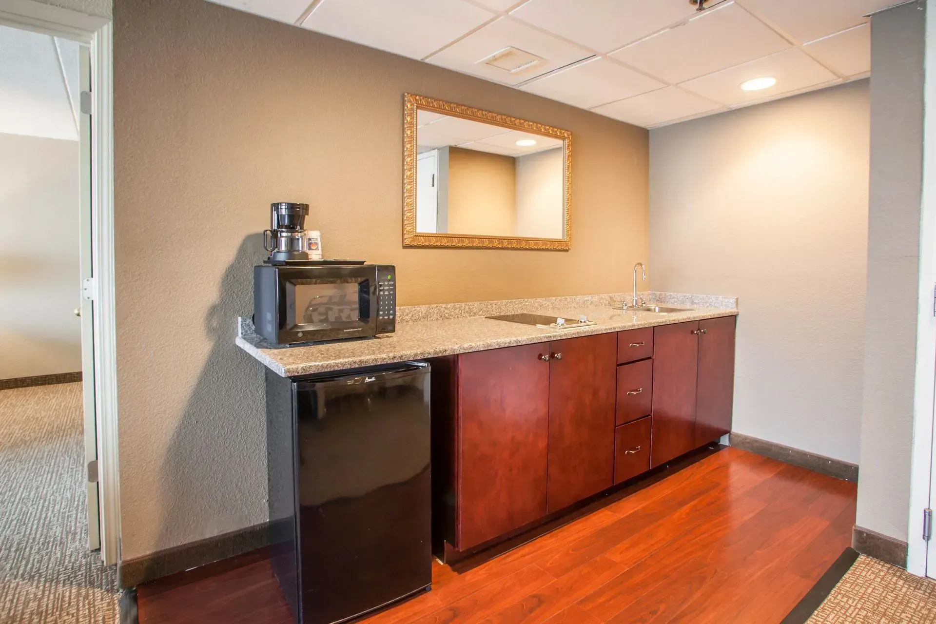 Office break room with sink, microwave, mini fridge, cabinets, and a wall mirror against beige walls and wood floor