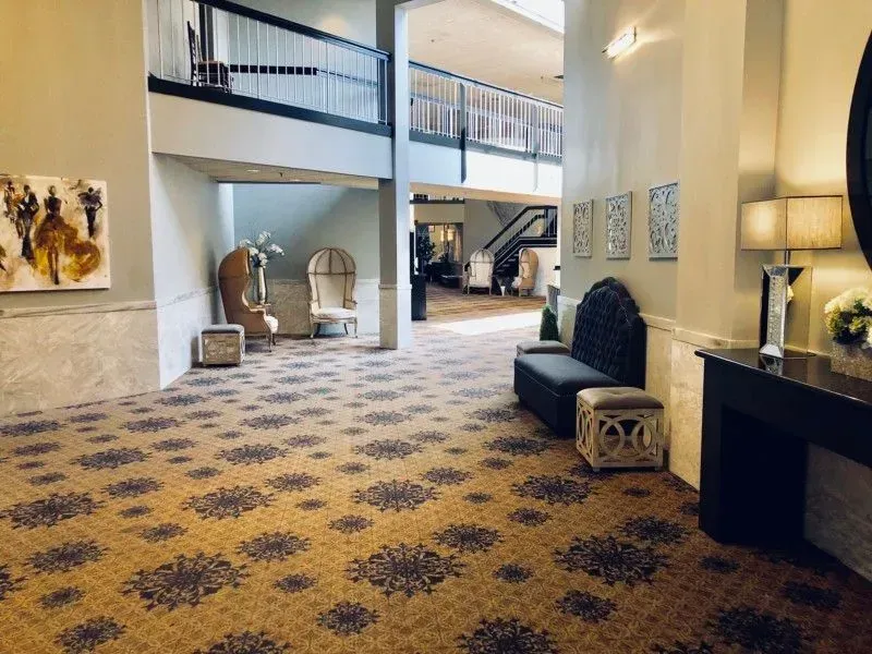 Hotel lobby with patterned carpet, seating, tall windows, and a staircase in a bright, open atrium
