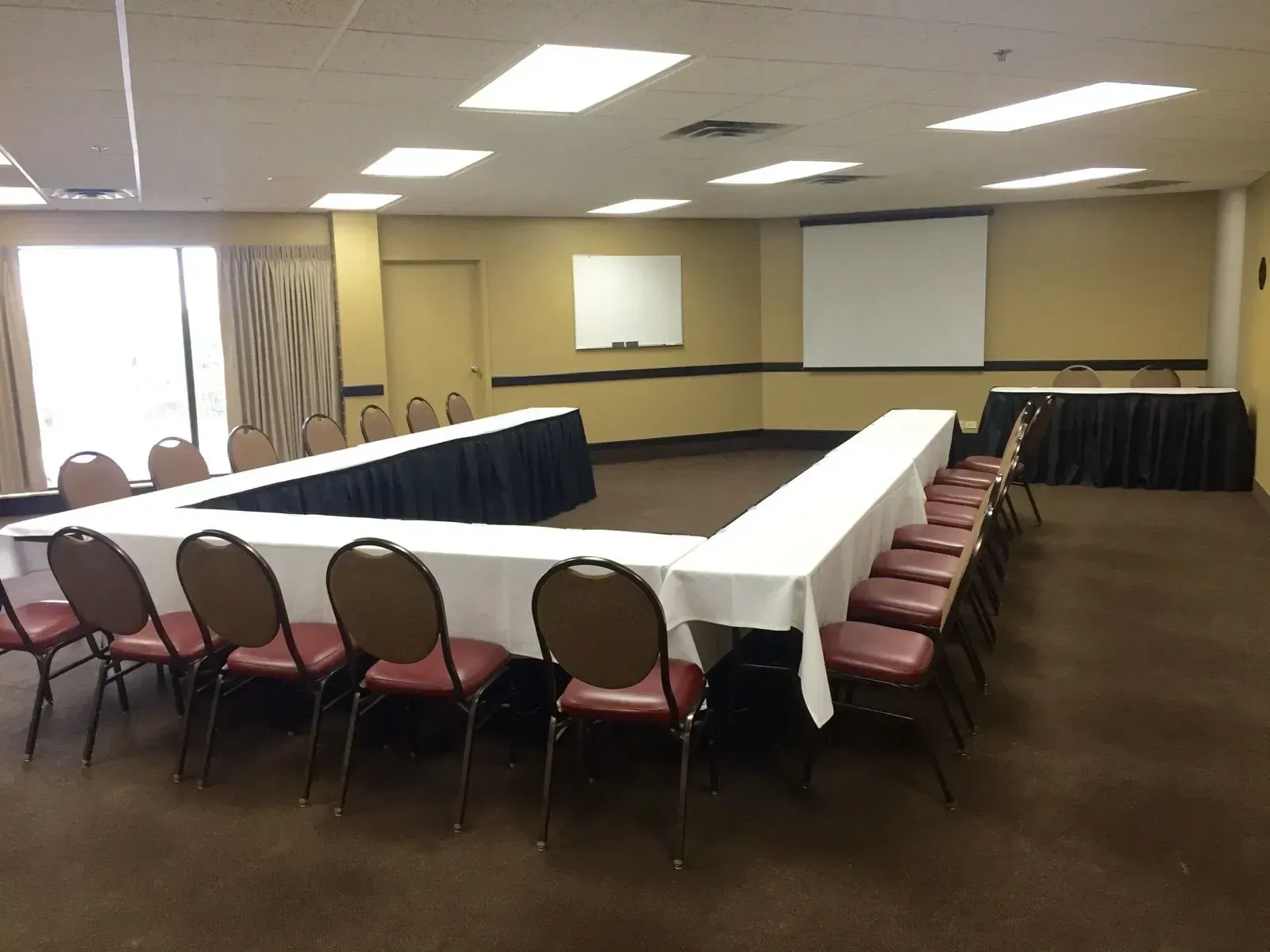 Empty conference room with U-shaped tables covered in white tablecloths and rows of red chairs, beige walls and carpet.