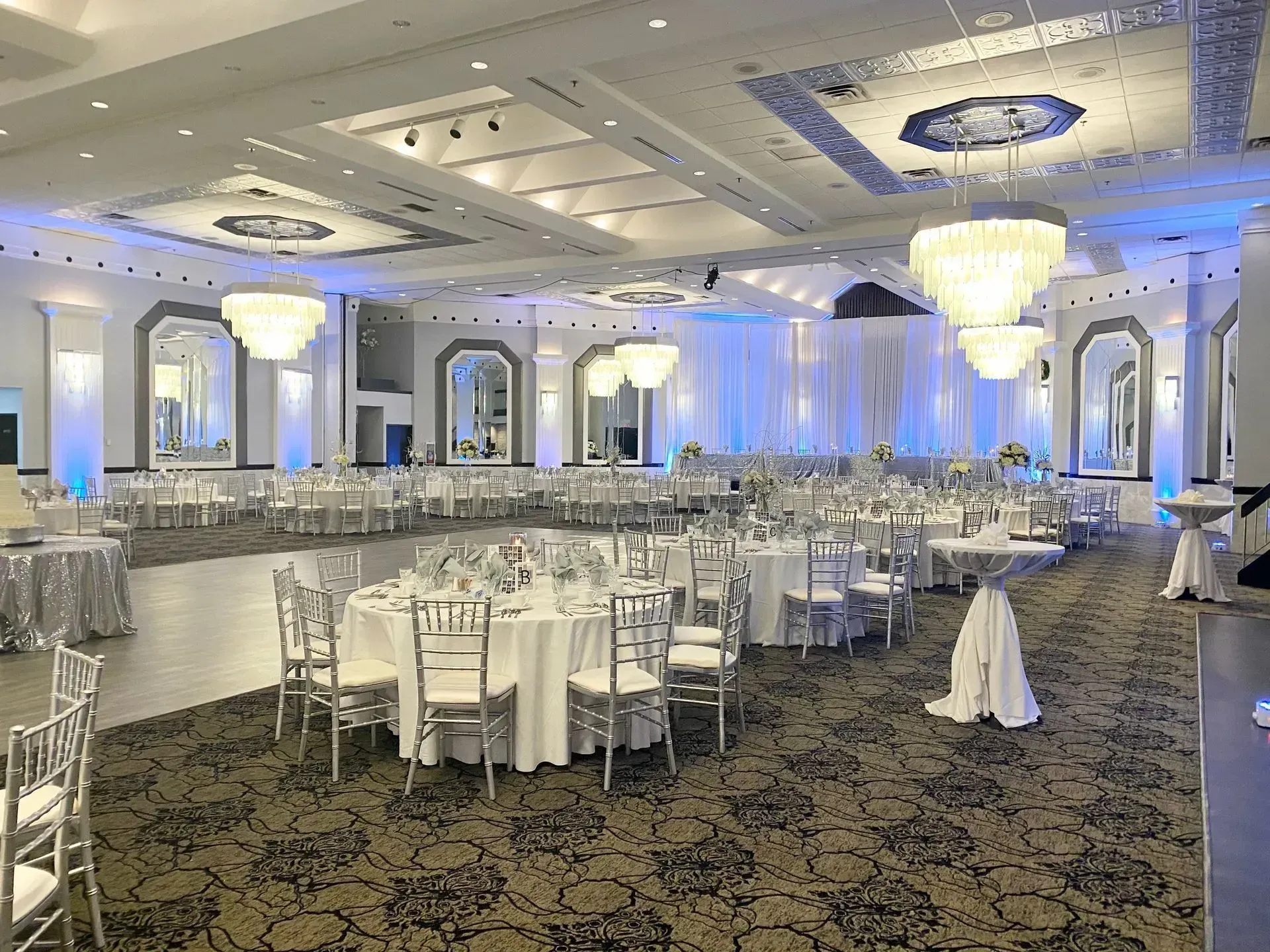 Elegant banquet hall with round tables, white chairs, chandeliers, and blue uplighting on the walls.