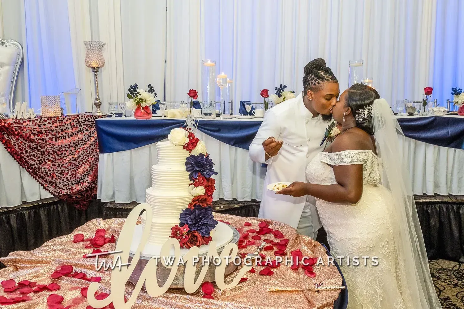 Bride and groom kiss beside a white tiered cake with red roses at an elegant wedding reception