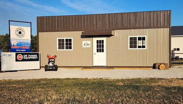 A beige metal building with a white door and dark brown trim stands on a gravel lot under a clear blue sky.