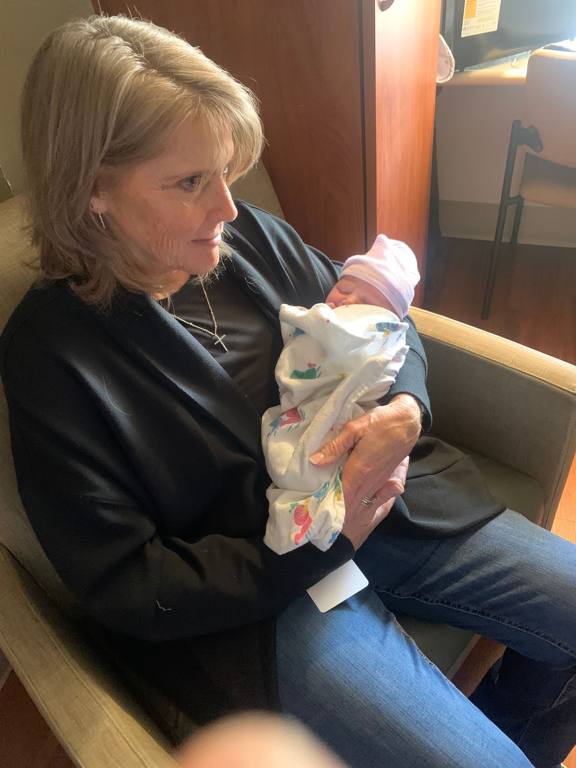 Woman in black cardigan holding a newborn baby wrapped in a blanket, seated indoors.