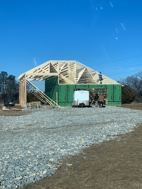 Construction of a building with wooden frame and green walls. Workers near a white trailer on gravel. Sunny day.