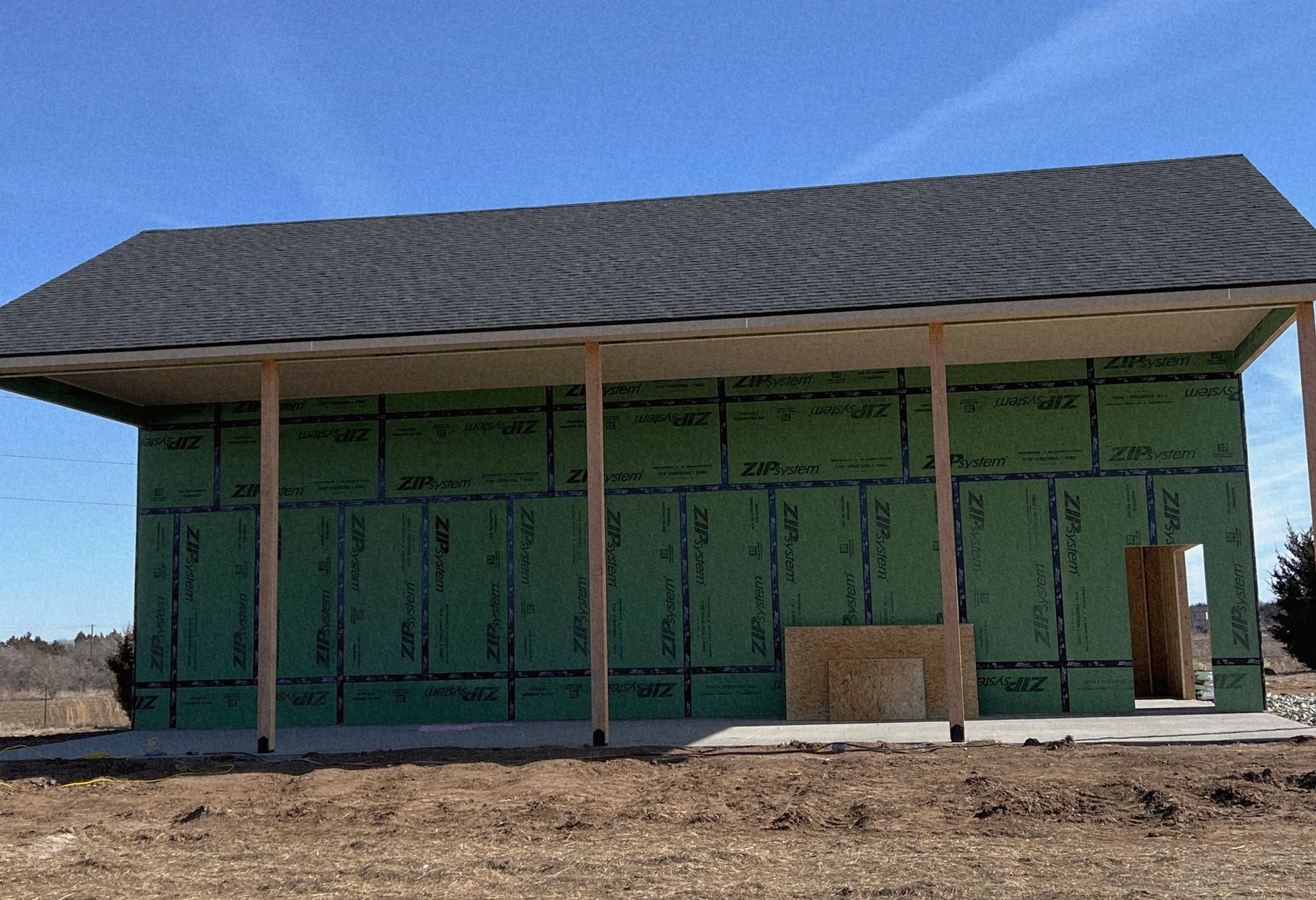 Unfinished building with green sheathing, a black roof, and wooden support beams on a concrete foundation, outdoors under a blue sky.