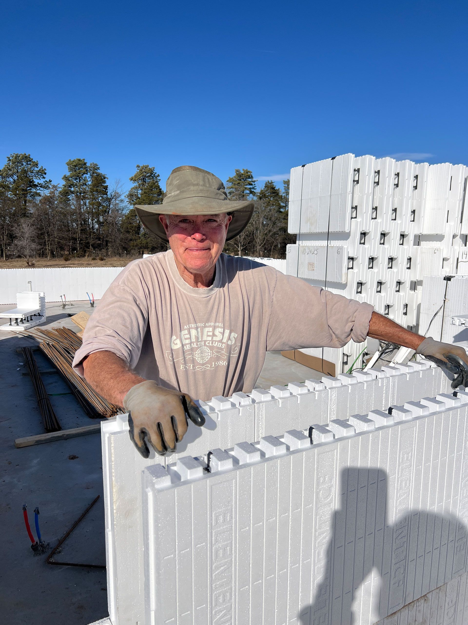 Man wearing a hat and gloves, building with white foam blocks outdoors under a blue sky.