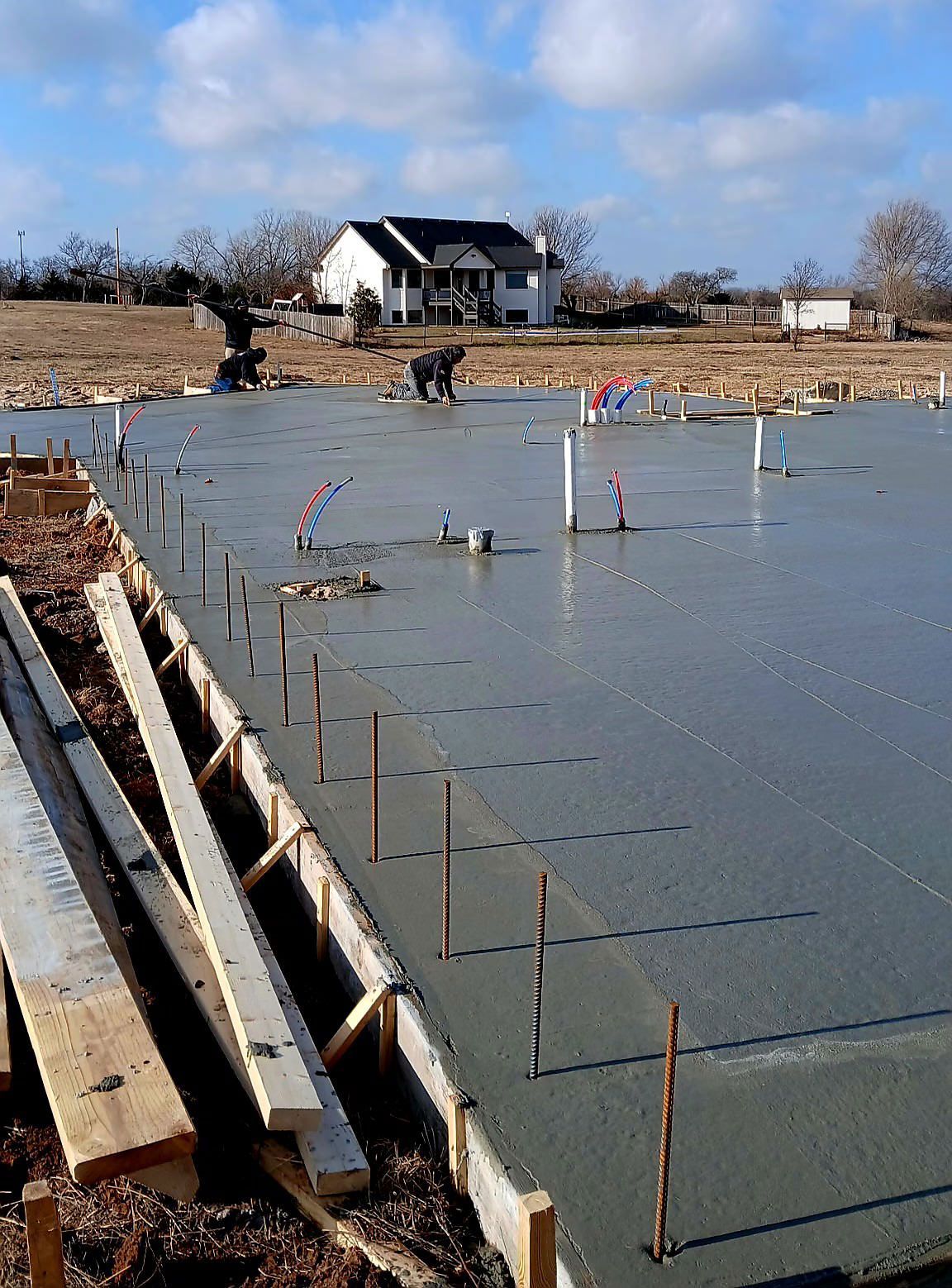 Newly poured concrete foundation with wooden frame, pipes, and stakes, under construction outdoors.