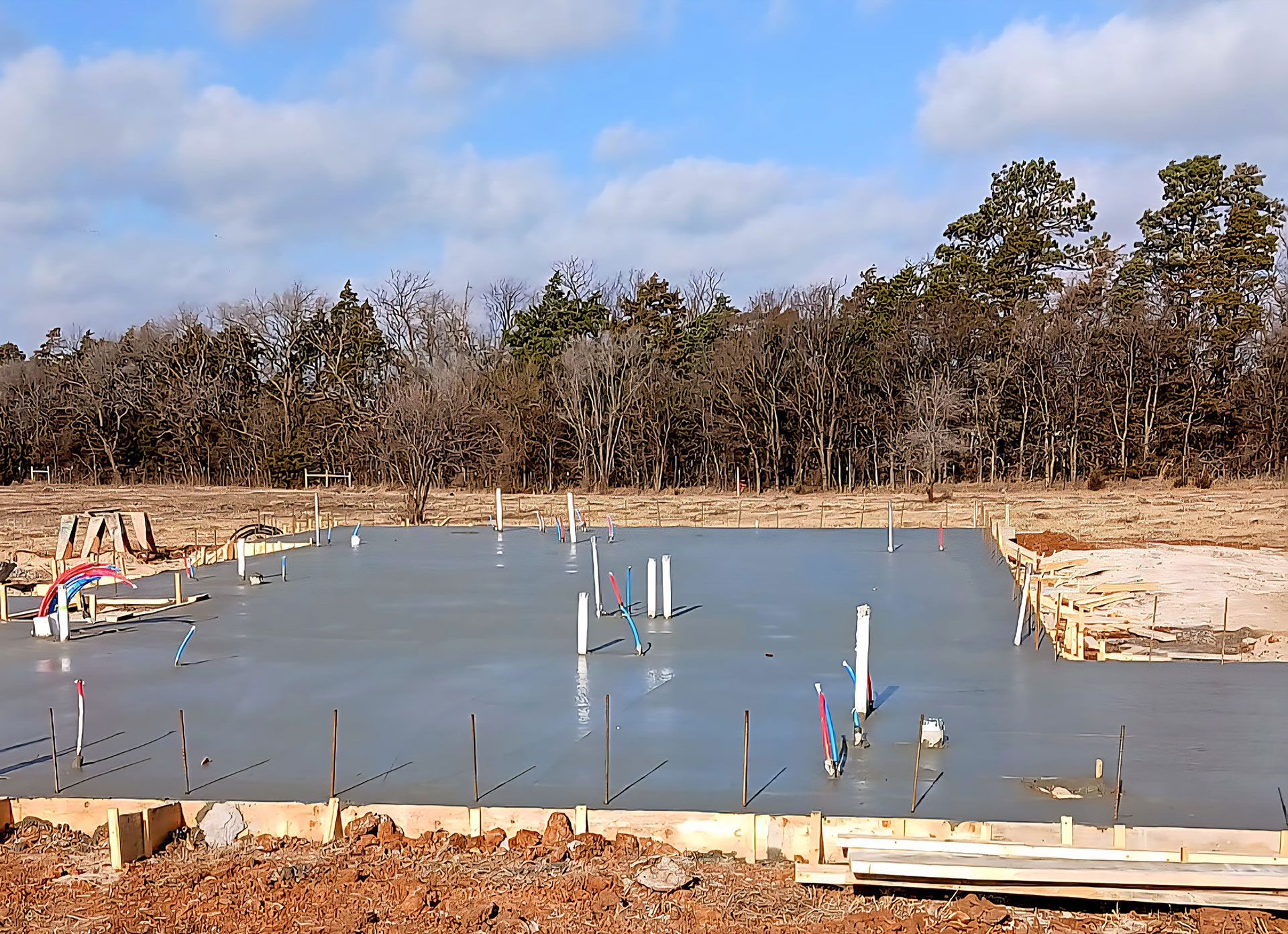 A freshly poured concrete foundation with plumbing pipes and wooden formwork, set in a construction site.