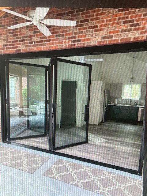 Black framed folding glass doors open to a kitchen. Brick walls and ceiling fan above, gray rugs and flooring.