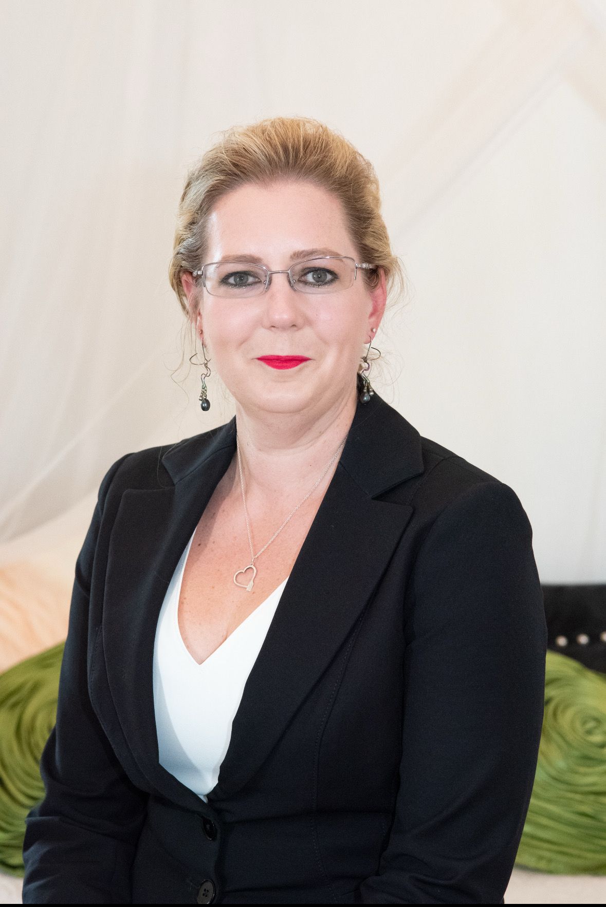 Woman in a black blazer and glasses, red lipstick, smiling in an office setting.