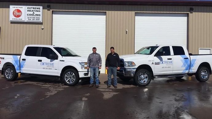 Two white utility trucks parked in front of a warehouse, with two people standing between them.