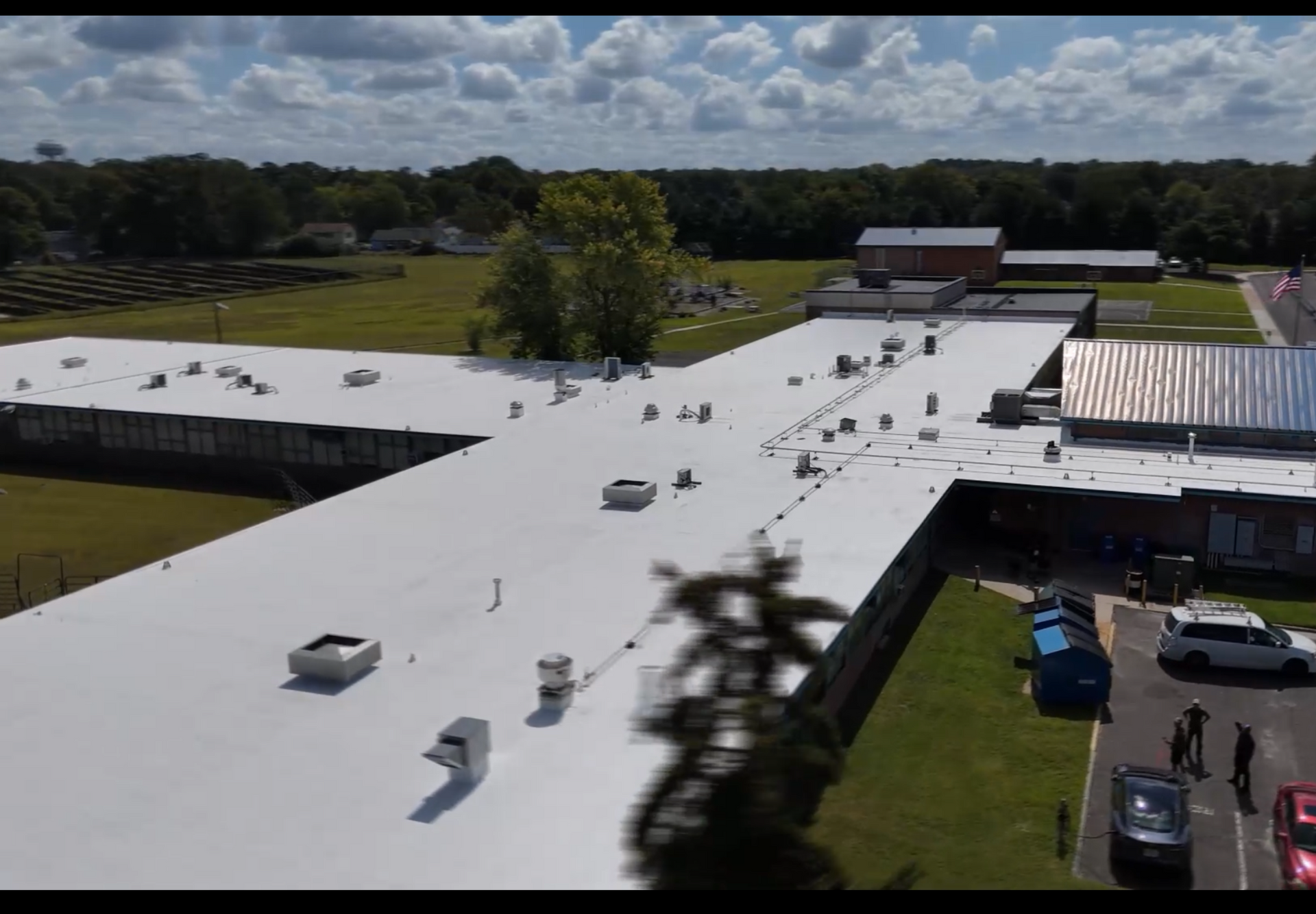 Aerial view of a white-roofed building with rooftop vents, parking lot, and trees under a cloudy sky