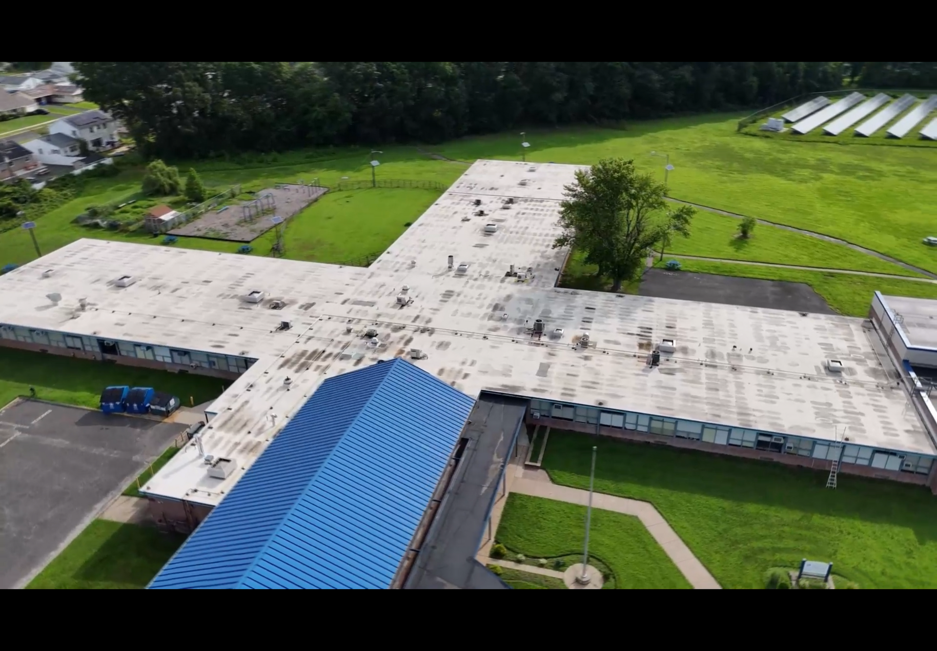 Aerial view of a large flat-roofed building with a blue entrance canopy surrounded by green lawns.