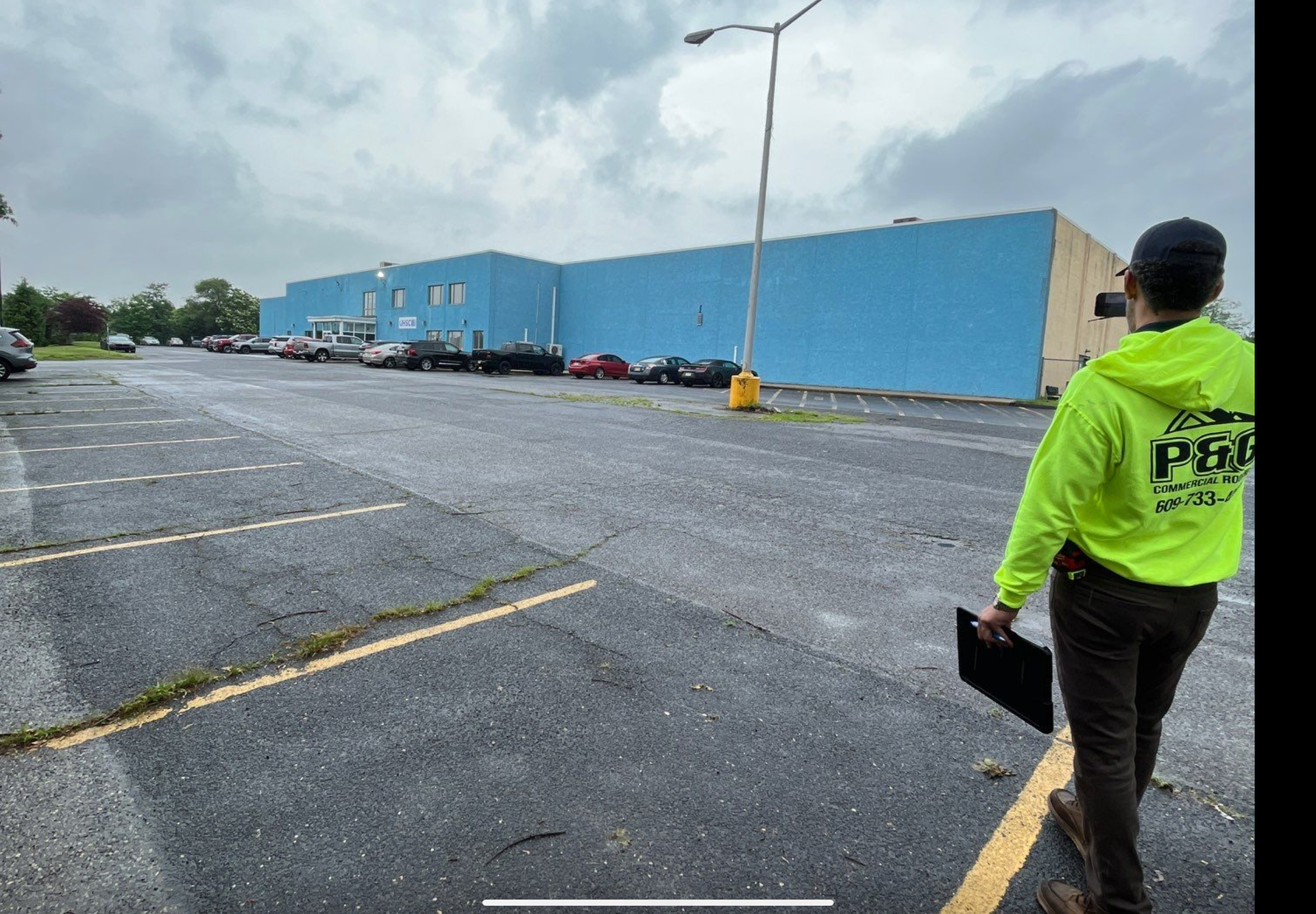 Person in a neon green jacket stands in a wet parking lot beside a large blue warehouse.