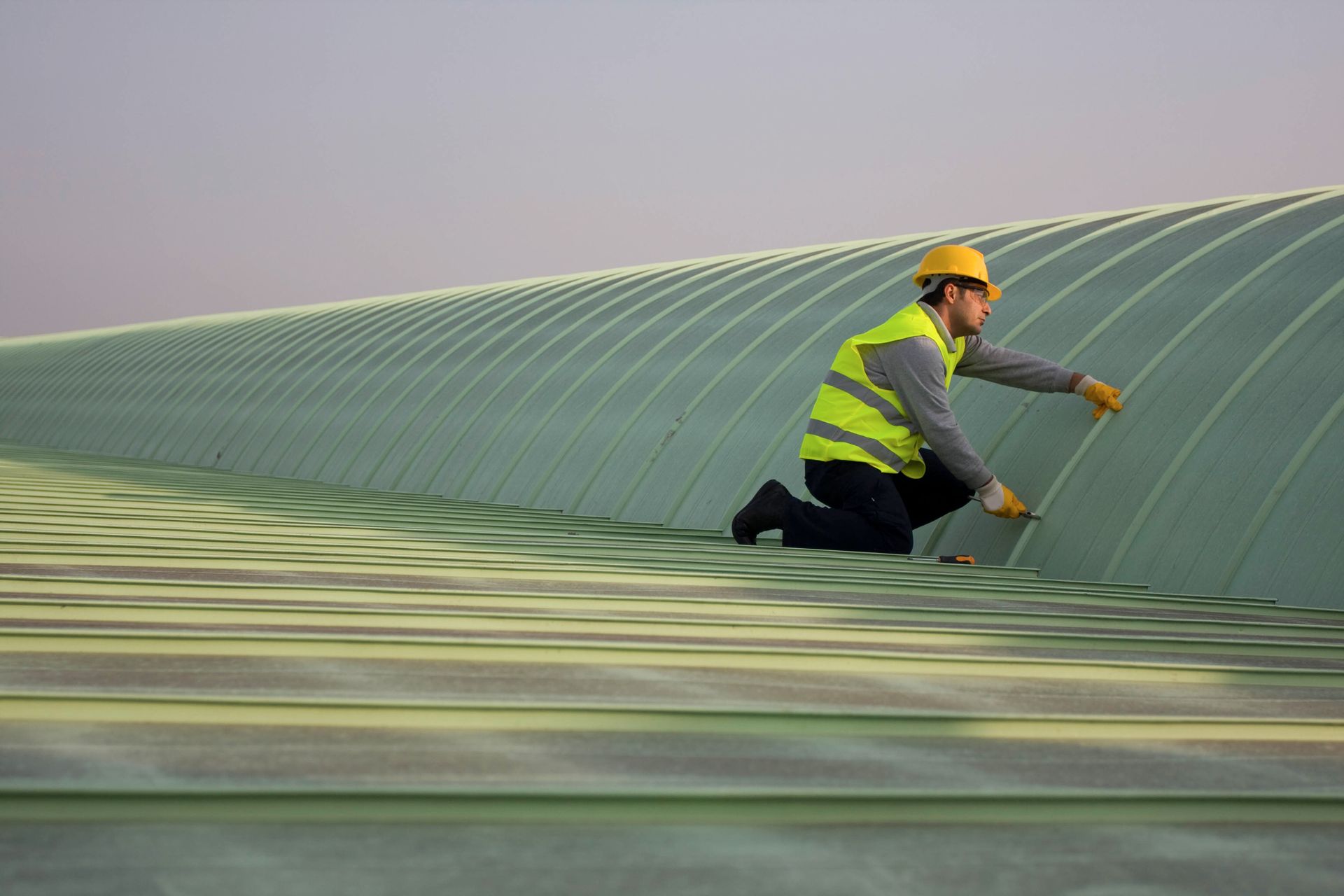 Construction worker in yellow vest and hard hat inspects a curved, green structure on a rooftop.