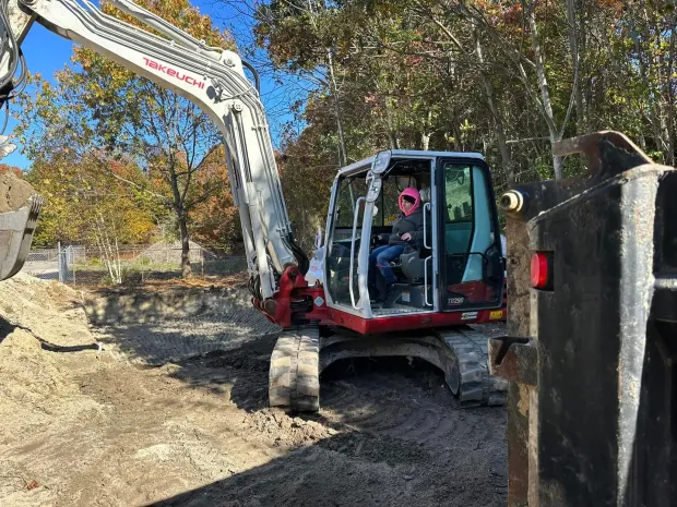 Excavator digging dirt beside a fence in an autumn wooded area, operator visible in cab.