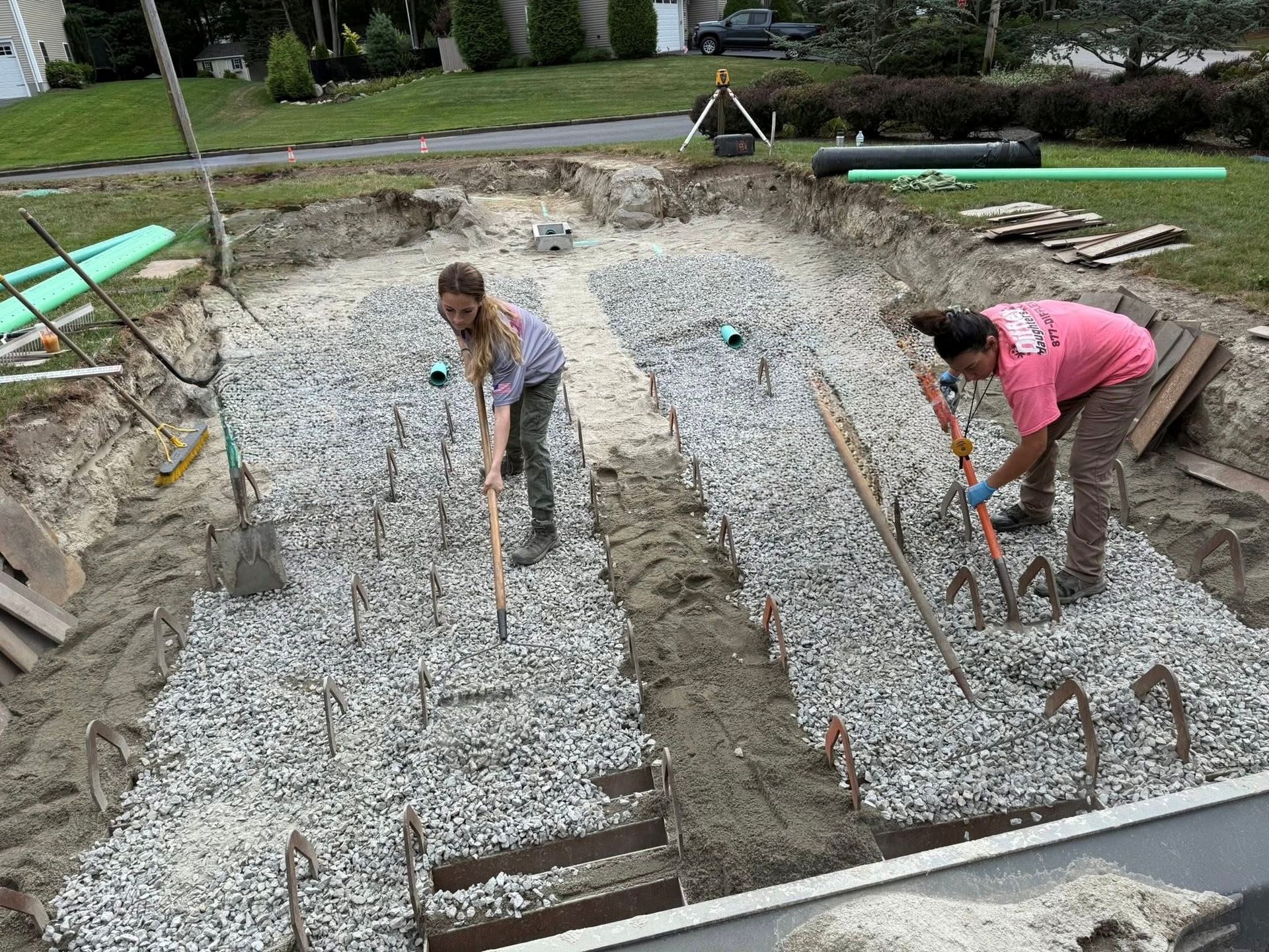 Two people using shovels in a gravel-covered construction pit with exposed rebar.