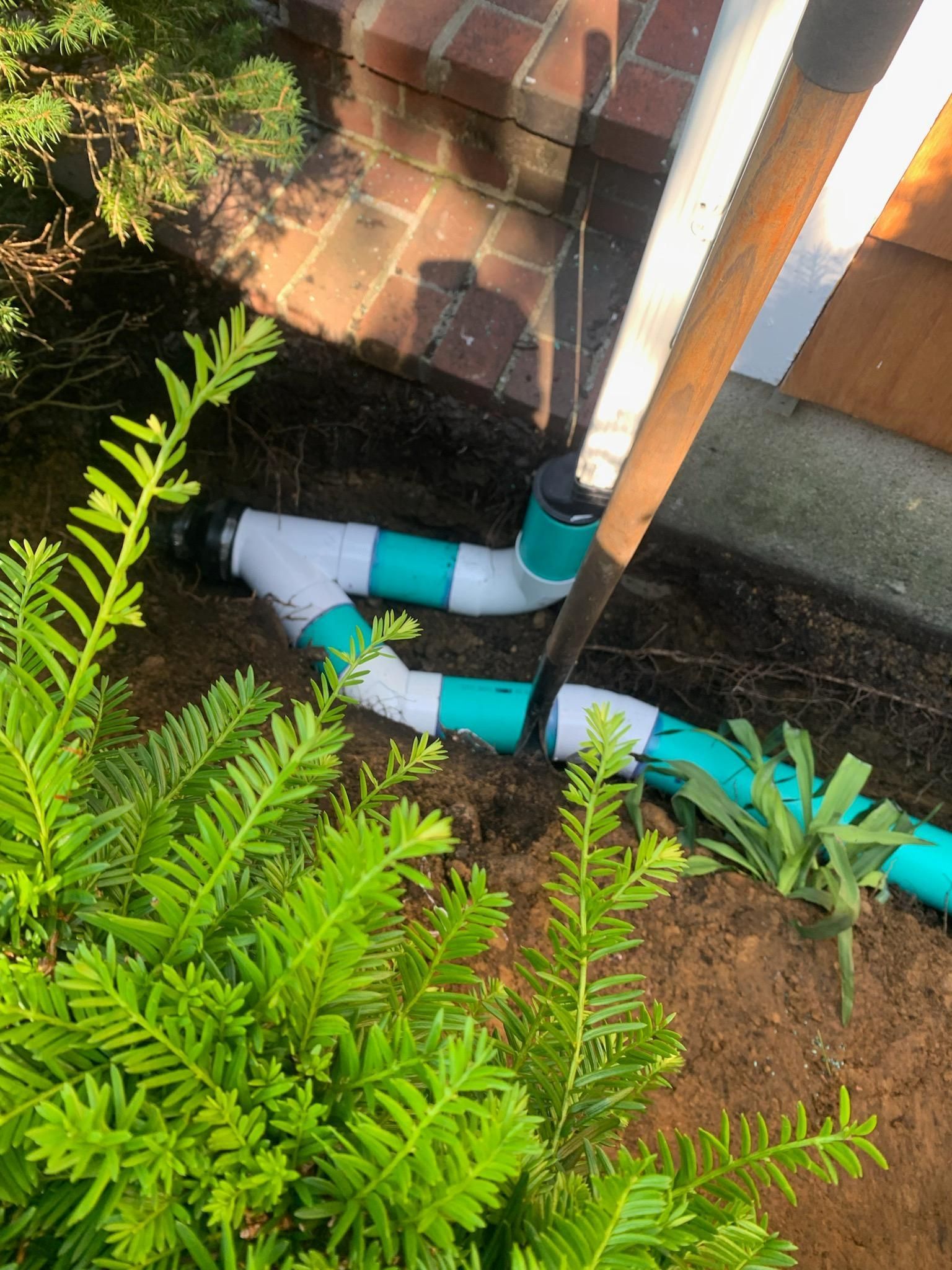 White PVC pipes with teal joints beside a brick wall, partly hidden by green plants and soil.