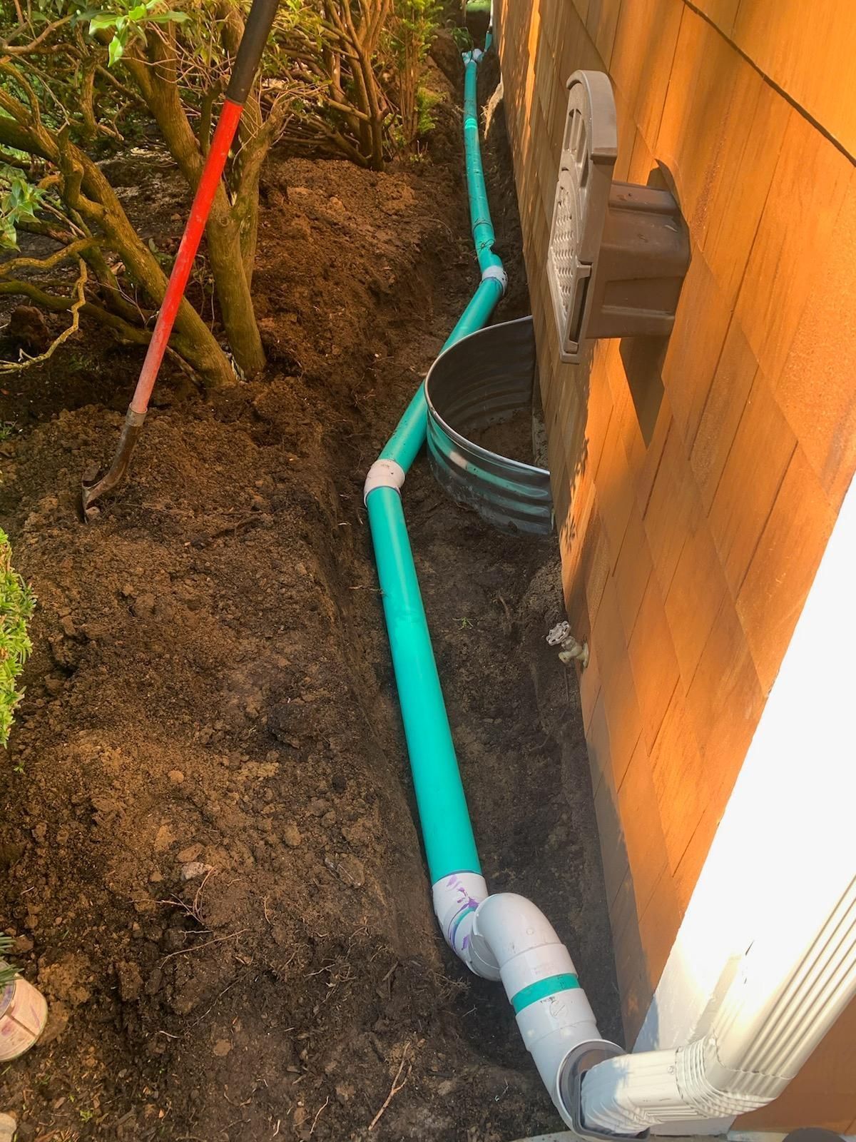 Trenched blue and white PVC pipes running along a dirt path beside a house wall and wooden fence.
