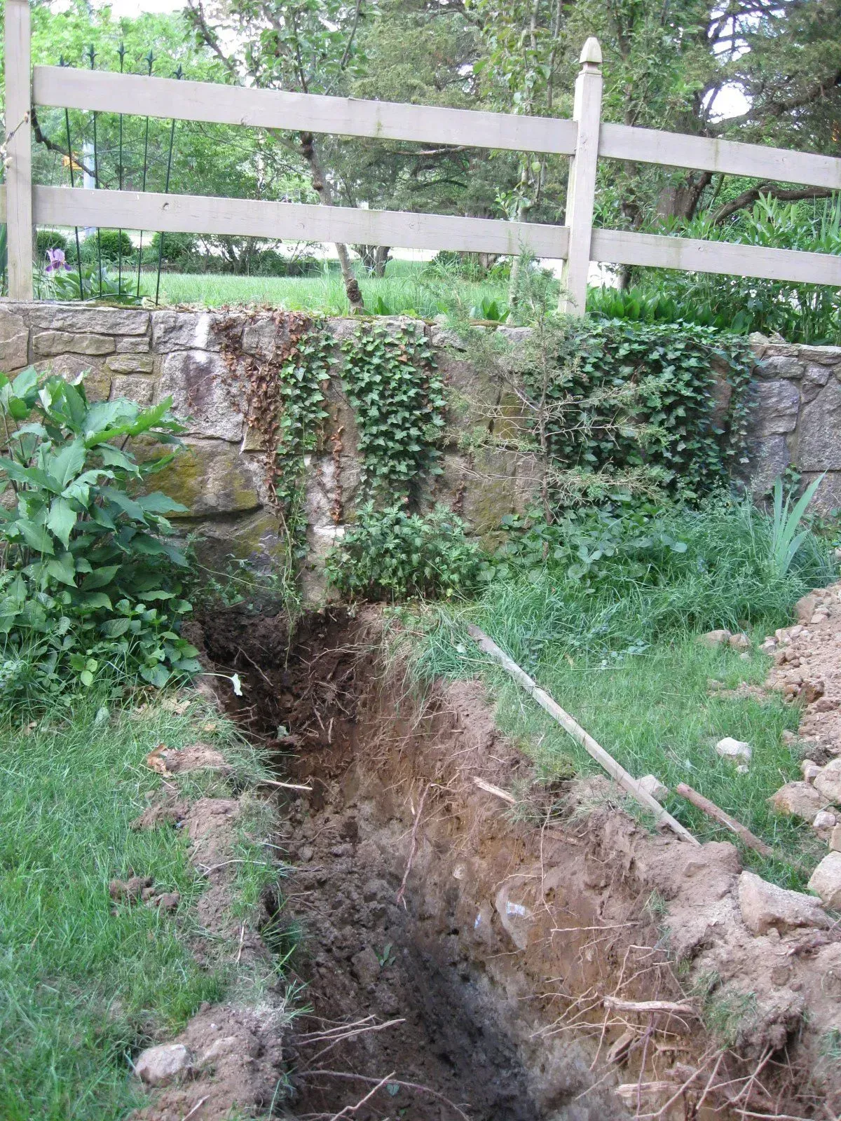 Backyard with a deep trench cut through grass beside a stone wall and white fence
