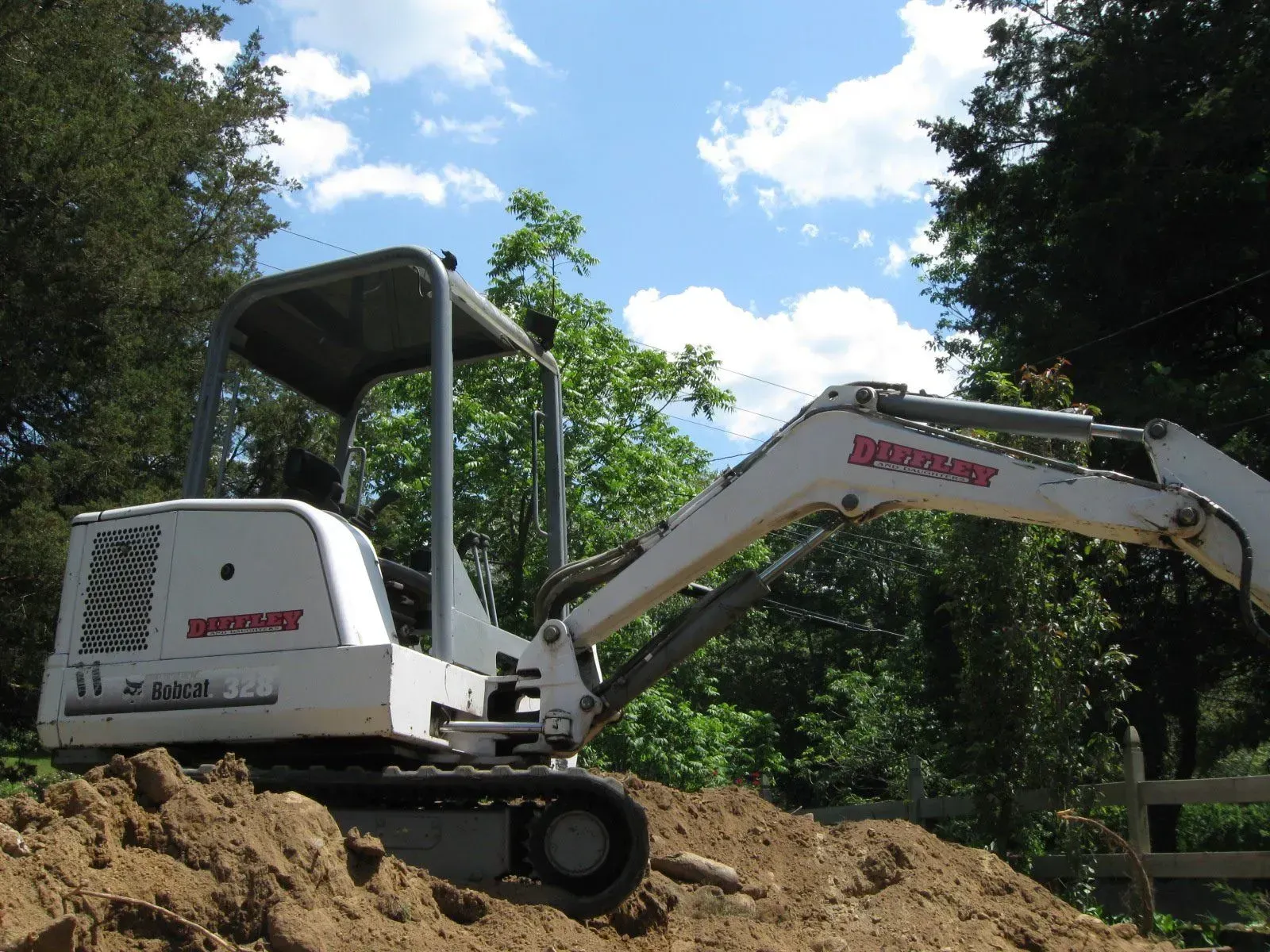 White excavator on a dirt mound at a wooded construction site under a blue sky