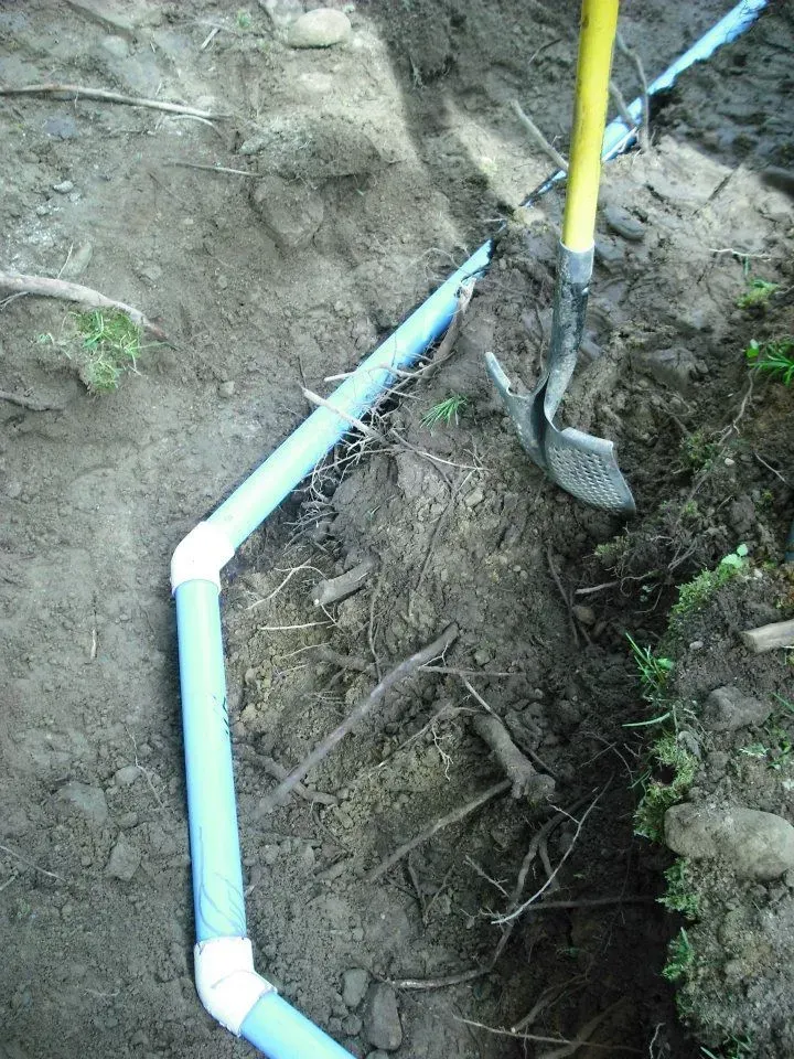 White PVC pipe laid in a dirt trench beside a shovel in an outdoor yard.
