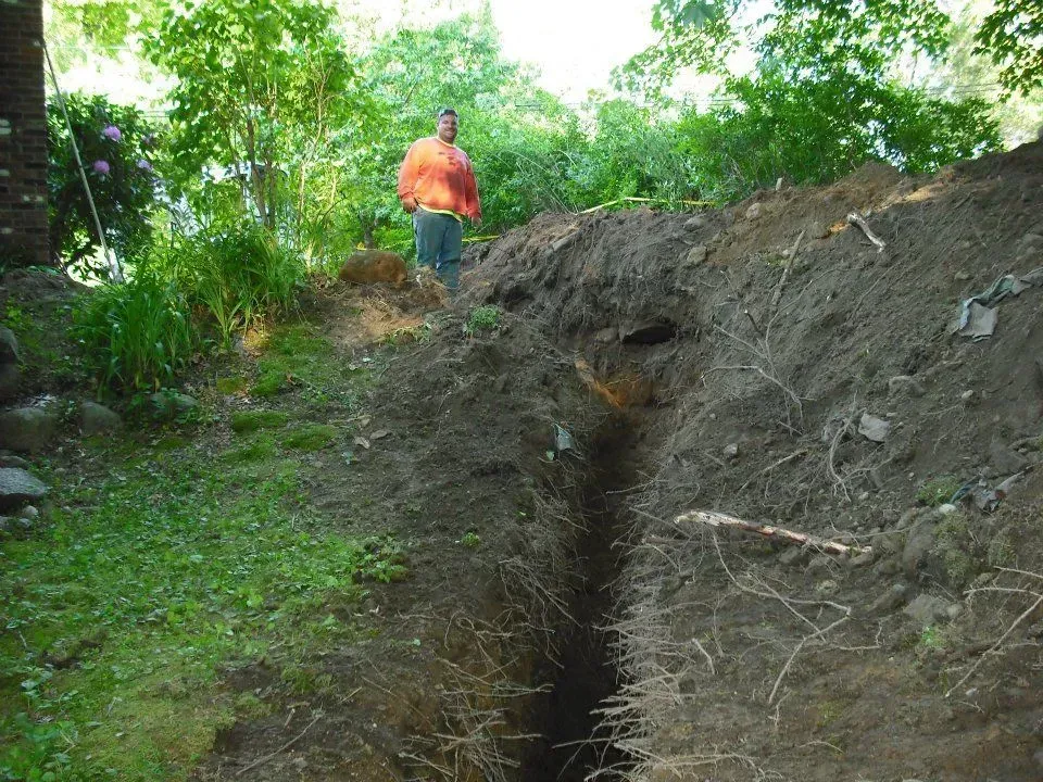 Person in orange jacket standing by a narrow trench in a wooded area.
