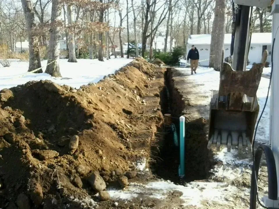 Trenched snowy sidewalk beside a large dirt pile, with a worker near an open utility ditch.