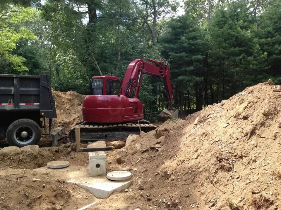 Red excavator at a dirt work site beside a dump truck and large soil pile in a wooded area
