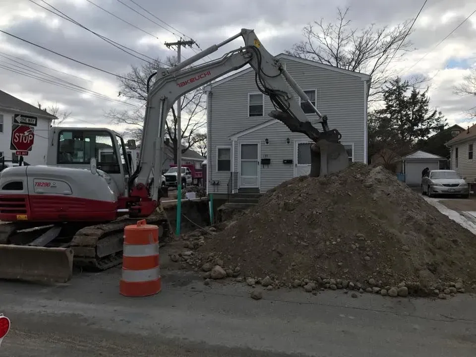 Excavator digging a dirt pile on a residential street beside a gray house and orange traffic barrel