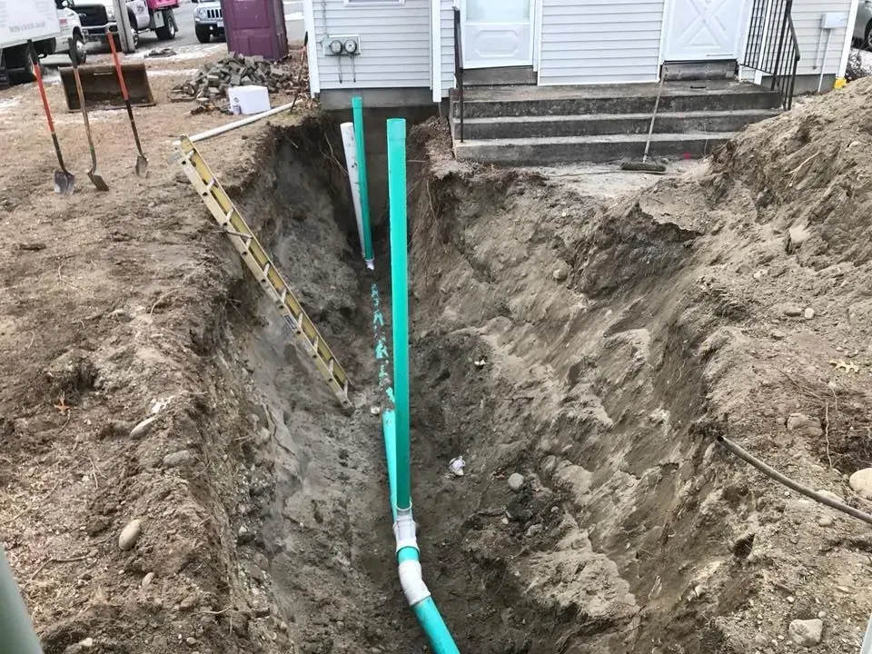Utility trench beside a house with exposed green pipes and a white PVC pipe running through the excavation.