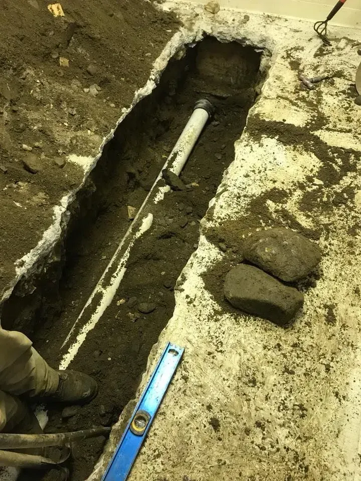 Trench with a white PVC pipe and blue level tool in muddy ground beside a worker’s boot.