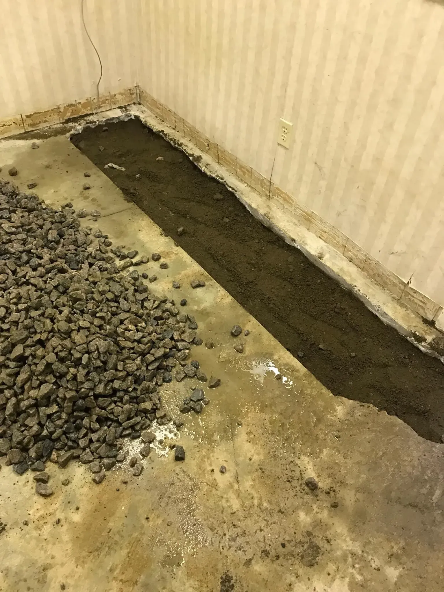Strip of dark soil beside a pile of gravel on a stained floor against a striped wall.