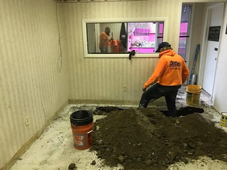 Worker in orange shirt kneels over a dirt mound during indoor room renovation.
