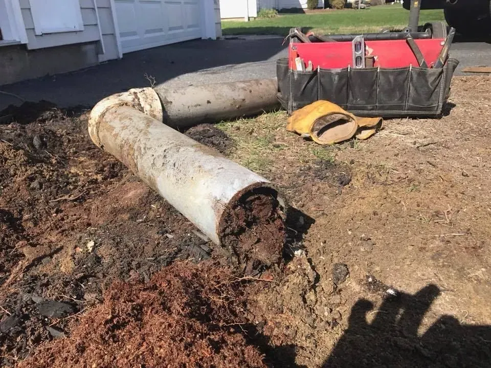 Large broken concrete pipe beside a house, with dirt piled nearby and a red trailer in the background