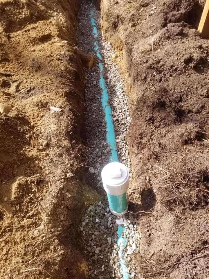 Trench with a blue pipe running through dirt, with a white capped PVC fitting in the foreground.