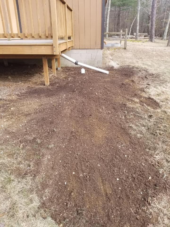 Brown dirt pile beside a wooden deck with a white drainage pipe in a wooded yard