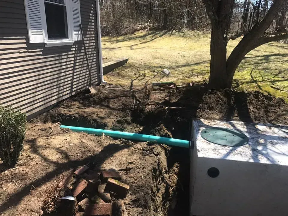 Backyard trench beside house with blue pipe, utility box, tree, and exposed soil and rocks