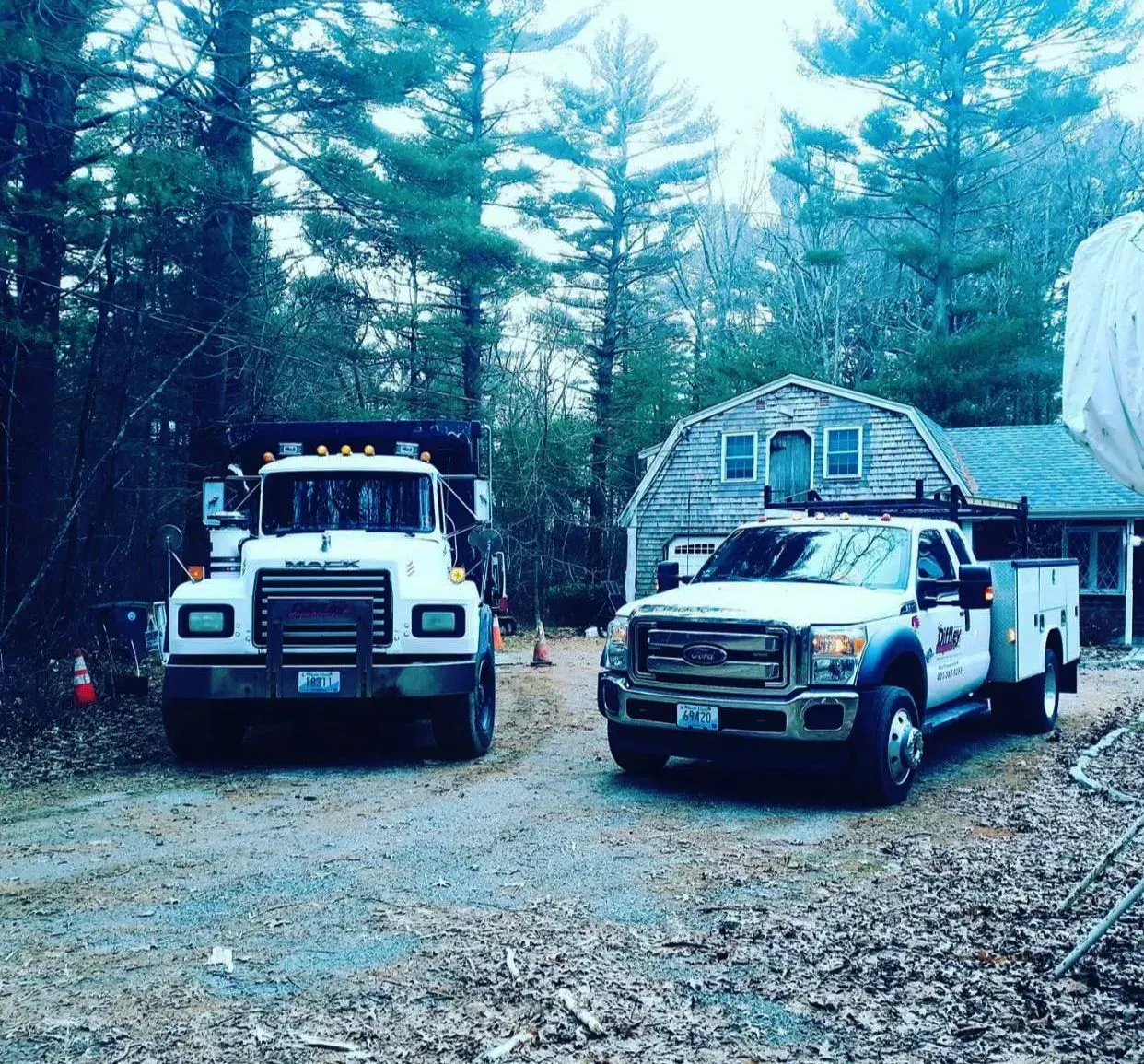 Two white utility trucks parked on a gravel driveway beside a small cabin in a wooded area