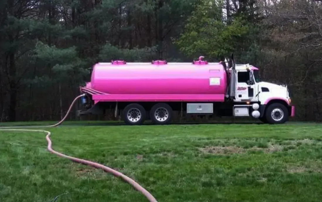Pink tanker truck parked on grass near a wooded area, with a hose trailing behind it
