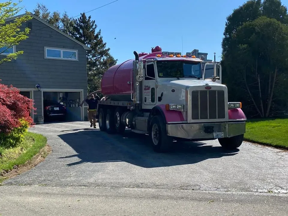 Pink tanker truck parked in a suburban driveway beside a house on a sunny day