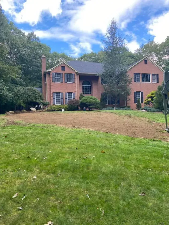 Brick two-story house with dark roof, bare front yard, and green lawn under a partly cloudy sky