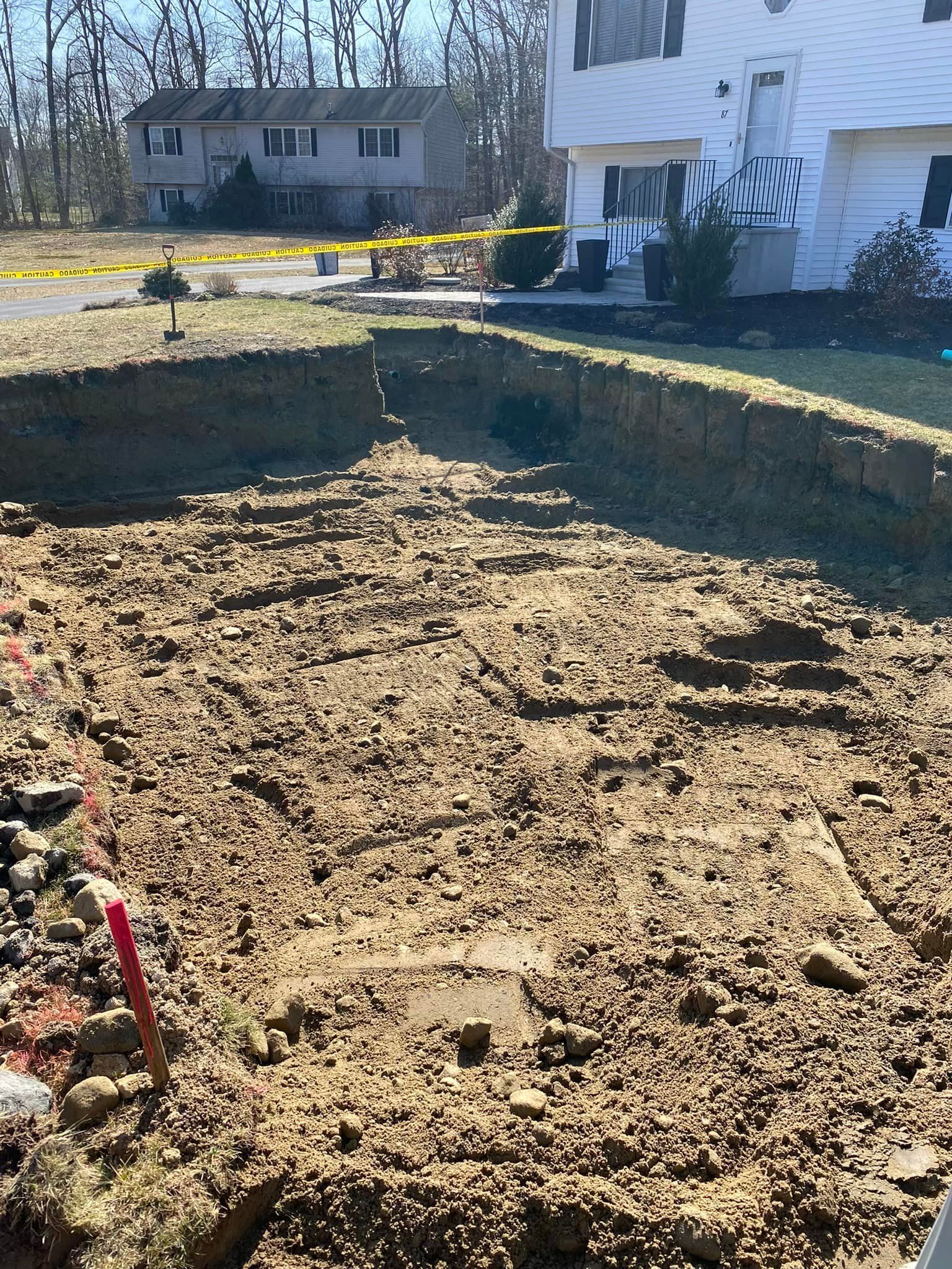 Excavated backyard with a large dirt pit beside a white house and yellow caution tape.